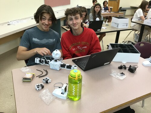 Two students are sitting on at a desk.  The student on the left has ear-length hair, is smiling and is wearing a grey t-shirt. The student on the right has a mullet, has an airpod in their right ear, is smiling and is wearing a red sweatshirt.  There is a