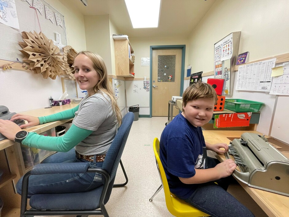 A young woman with long blonde hair and a young boy with short brown hair smile at the camera.  She is wearing jeans with a grey t-shirt with green sleeves and jeans.  He is wearing a blue t-shirt with jeans.  Both are sitting in a small office at Perkins