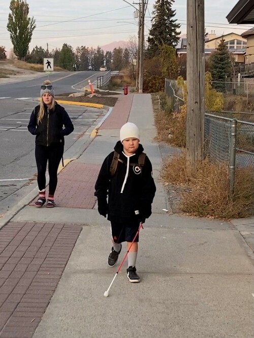 A young male student is walking on a street with a white cane.  he is wearing a white toque, blue hoodie, black shorts, grey socks and black sneakers.  In the background a middle aged female follows him