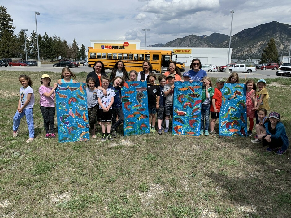 A class of students holding four blue panels with salmon pained on them.  Bus parked behind them.