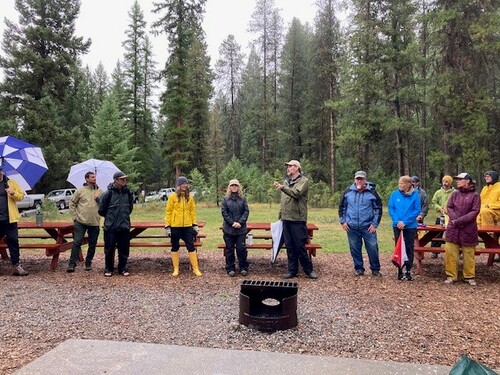 11 adults in a park wearing rain gear and with umbrellas.  There are evergreen trees in the background and a firepit in ghe foreground.